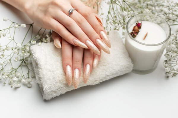 Tipos de alongamentos de unhas - mão com unhas alongadas em fundo trabalhado com toalha e copo tons pastéis